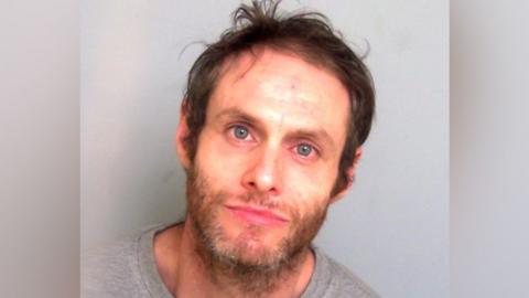 A man with longish brown hair stares at the camera. He has blue eyes which look slightly red and a scruffy beard. He is wearing a grey top and stands against a grey background.