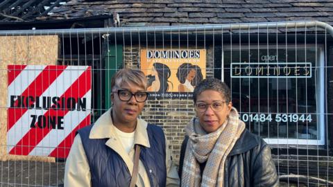 Two women with short hair stand outside fencing which guards their business premesis, which had been fire damaged.