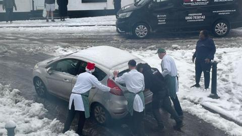 Men in white shirts and green aprons and santa hats  pushing car in snow 
