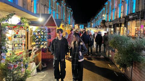 Man and women walking through a Christmas market at night, with stalls selling Christmas gifts and Christmas trees and lights running along the side.