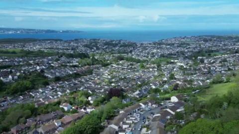A drone photograph of an area of Brixham which shows lots of houses, roads and some trees as well as the sea further away. 