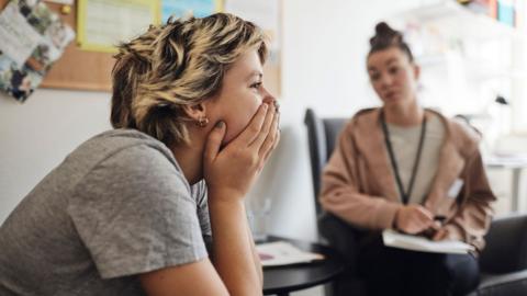 Stock image shows a young person with short blonde hair with their hands over their mouth looking pensively away from camera while a counsellor in the background speaks and writes on a piece of paper.
