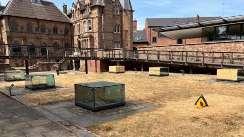 The image shows six glass display cases, all of which have been damaged and had some of their glass panels smashed. The damaged panels have been boarded over. In the background is the historic Blue Coat School and to the right, the Priory Visitors' Centre.