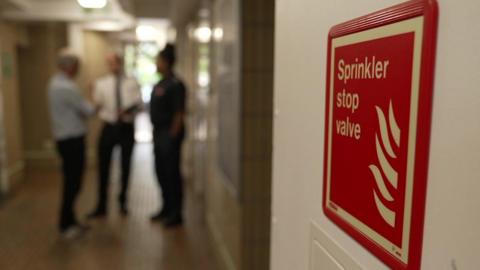 A sprinkler sign in display in a building is in the foreground with London Fire Brigade staff in the background