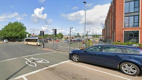 A crossroads, with a blue car in a lane in the foreground behind traffic lights on red. A bus going in the opposite direction is moving across the junction. 