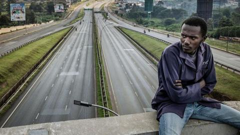 A stranded worker in dark blue top and blue jeans sits on a bridge overlooking an empty Thika highway, one of Kenya's main highways