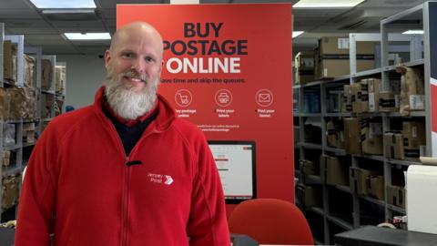 Chris wearing his red Jersey Post fleece jacket standing in front of the counter at Jersey Post headquarters. A sign behind him says "Buy postage online: Save time and skip the queues."