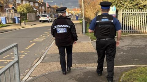 The backs of two police officers seen patrolling along a pavement in a residential area. It looks as though they are walking past the gate of a school or nursery. They wear black police uniform and hats.