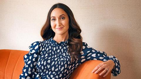 Eve Simmons, sporting long brown hair and a blue dress, is smiling while sitting on an orange sofa