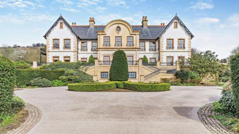 The entrance to a mansion. The property is a beige and white colour with perfectly trimmed bushes in front of the house. A circular driveway can be seen looping around the entrance to the property.