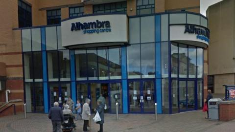 The front of the Alhambra Shopping Centre with a group of about nine people standing in front of it on a brick concourse