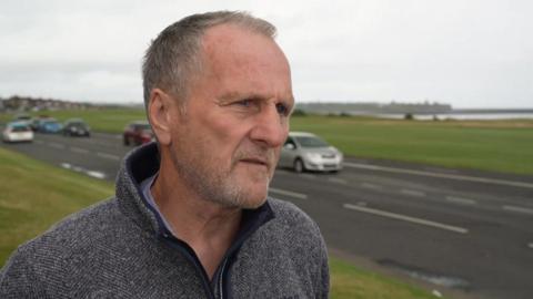 Dave Dixon, campaigner, with short grey hair, wearing a grey fleece, looks to the right as he stands on a grass field, with a road with cars driving past next to him. 