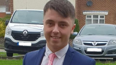 Head and shoulder shot of Connor Brown, a smiling young man with short brown hair. He is wearing a dark blue suit and a pink tie. Behind him are a van and a car parked on a grassed area.