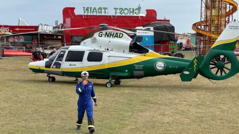 An air ambulance is landed on a grassed area in front of several fairground rides. A member of the emergency services can be seen in the foreground. He is wearing a blue uniform and white helmet.