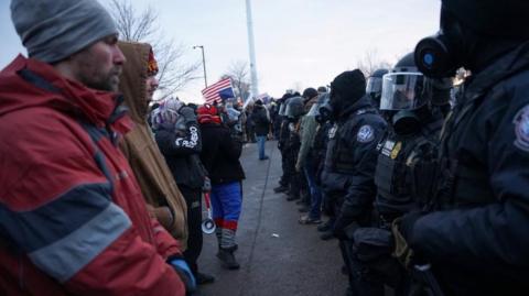 Demonstrators stand in a row in front of members of US Customs and Border Protection and other law enforcement officials, near the Bishop Henry Whipple Federal Building. Federal agents are dressed in black and wearing gas masks that cover their face. Demonstrators are wearing plain clothes. Some in the back are waving an upside-down American flag.