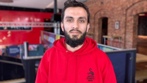 A young man with short dark hair, beard and moustache is standing in a brick-built student union, wearing a red University of Lincoln hoodie and staring into the camera. 