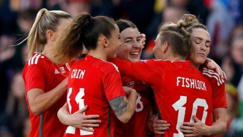 Wales' Angharad James celebrates scoring a second goal with teammates. She is smiling and being hugged by four other players in their red Wales shirts, including Fishlock and Cain. The background is blurred. 
