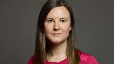 Official portrait of Labour MP, Maya Ellis. She has long, straight dark hair and a red top. She is standing in front of a plain dark grey background
