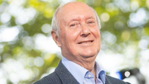 Sir Kenneth Calman in open necked blue shirt and blue checked jacket is smiling  in front of a de-focussed background of trees and sky.