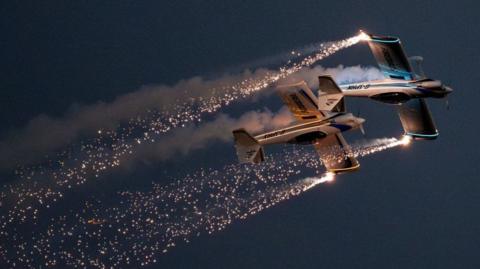 Two Firebirds planes fly through a dark sky with sparks coming from their tails.