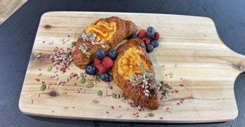 Two Croissants on a wooden board with a crème brûlée filling, dressed with blueberries and raspberries and mixed seeds.