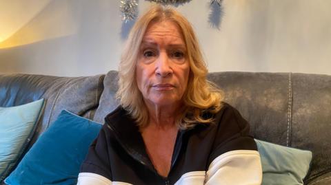 Sue Rombach has blonde hair to her shoulders and sits on a grey sofa in front of a white wall with blue and green cushions on the sofa. She has a black top with a white horizontal stripe on it.