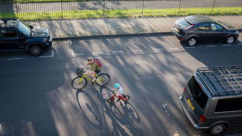 A woman guides a small child while riding their bikes along a minor road. There are cars parked along the side of the road and a fence visible on one side of the pavement.