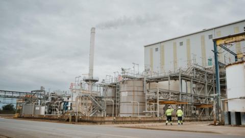 The titanium dioxide (TiO2) pigment production facility at Greatham, in Hartlepool. Two men in high viz jackets and hard hats walk towards industrial buildings. There is a large smoking chimney on the left, with tanks and several sets of external stairs visible on the right.