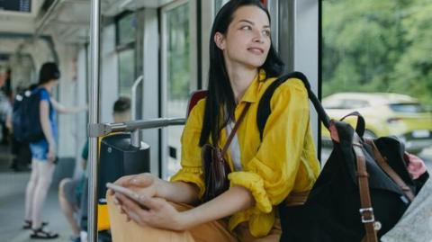 A woman sits on a bus and looks out of the window. She is wearing a yellow shirt and has a black shoulder bag and a phone in her hand.