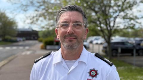 A man with grey hair and silver-rimmed glasses is standing at the entrance to Lincoln County Hospital. He has a stubble beard and is wearing a white shirt with black lapels, and a fire and rescue badge.