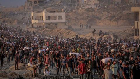 large crowds, some carrying sacks, walk past ruined buildings in Gaza