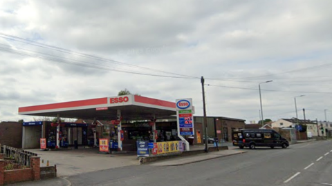 A picture of a petrol station forecourt with the word Esso above the station with a wall to the left of the forecourt and a brick building to the right of the petrol station