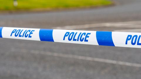 A section of a road cordoned off with blue and white police tape marked 'POLICE' in bold letters. The scene is outdoors, featuring a curved road and grassy areas.