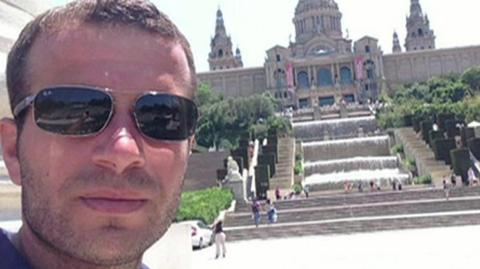 Albert Xhediku poses for a selfie in front of the Palau Nacional in Barcelona, a palace with domed turrets behind a cascading water feature and steep steps down to where he is standing. Mr Xhediku has short, brown hair and a close-cropped beard and is wearing sunglasses.
