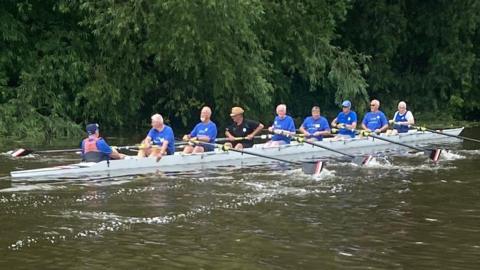 A group of eight men in blue t-shirts rowing. There is a cox at the back of the boat. 