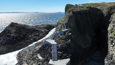Staffa's new staircase in sunshine. The steps are made of concrete and go up the side of a steep sea cliff. Sunshine sparkles on the sea behind the island. 