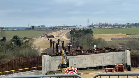 A bridge being built. There are metal pillars where the structure will be placed. Beyond it is a road under construction.