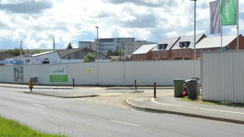 Lagan tvs site on Marlborough Road. A fence can be seen around the development and some new houses can be seen in the distance behind it. There are also flags for Lagan tvs along the path. 