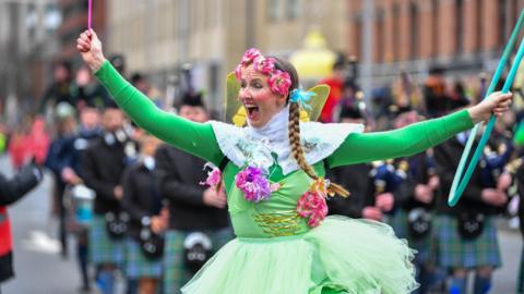 A woman dressed in a green fairy outfit. She has pink flowers around her head and holding a wand in one hand and a hoola-hoop in the other.