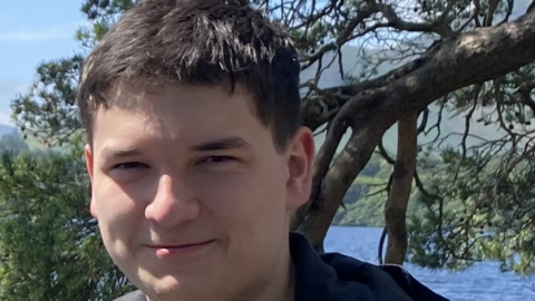 Tom Dingle smiling for a photograph. He has short dark brown/ black hair and is standing in front of a tree and a body of water.