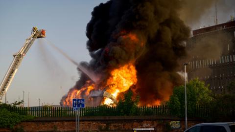 A large fire engulfs a building, with significant flames rising from the ground, creating a large black cloud of smoke, while a jet of water from an aerial hose works to fight the fire.