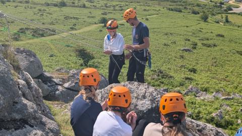 Three girls in orange helmets watch their friend abseil down a rock face with the help of an outdoor instructor.