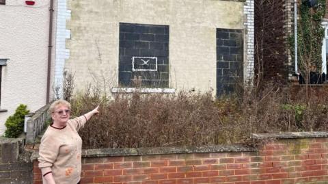 Kathleen Wilson, wearing a beige, is standing outside a badly overgrown garden in front of a derelict house. She is pointing at the house which has concrete blocks in the windows and doorway. There is a waist-high brick wall between her and the property.