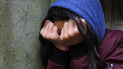 Library photo of a woman with a red jacket and blue hood, with her head in her hands. She has dark hair and is leaning against a concrete wall.
