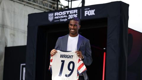 Haji Wright wearing a blue suit jacket and white t-shirt poses with his USA shirt at the event to reveal the USA's World Cup squad