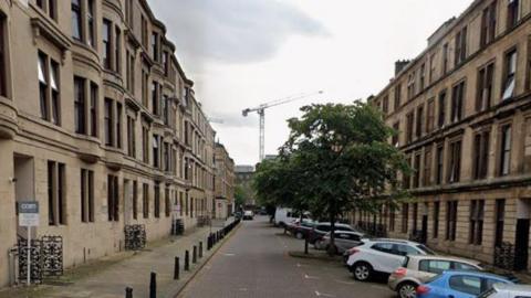 A long street of blonde sandstone tenements with some cars parked in front of them.
