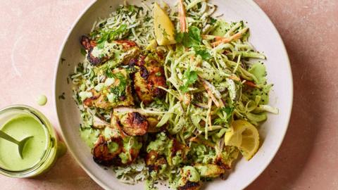 A white plate on a mottled pink background filled with slaw, herby rice, browned chunks of chicken and squeezed wedges of lemon