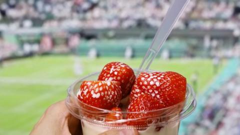 A person holds a pottle of strawberries and cream with a tennis court in the background
