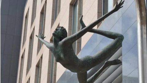 A naked girl made out of metal with a green tinge has her arms outstretched and her legs thrown back behind her. She appears to be floating.