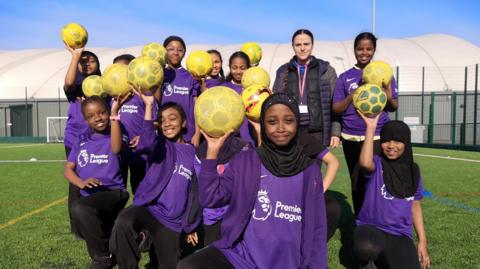 A girls' football team wearing purple Premier League shirts and jumpers. They hold yellow footballs in the air and stand on a football pitch. Some of the girls wear headscarves. They stand with their PE teacher, who wears a black coat.